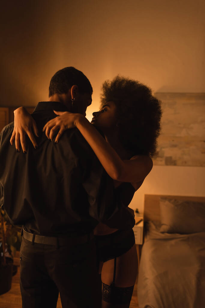 curly and sexy woman hugging african american boyfriend in black shirt in dark room with lighting - Photo, image