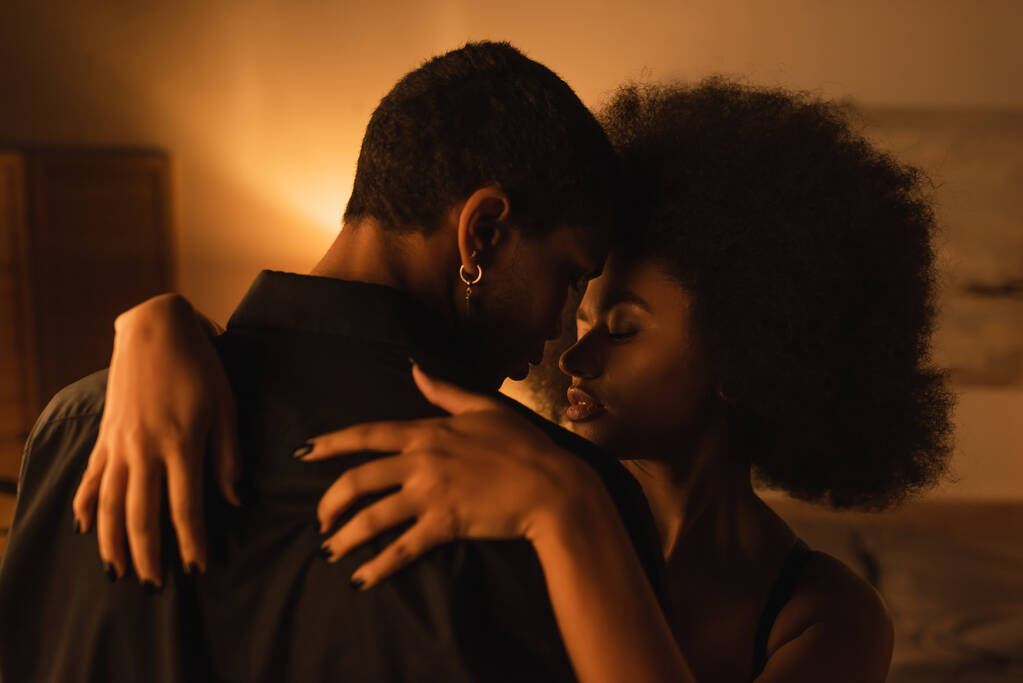 joven afroamericana americana mujer con pelo rizado abrazando joven novio en negro camisa en oscuro dormitorio - Foto, imagen