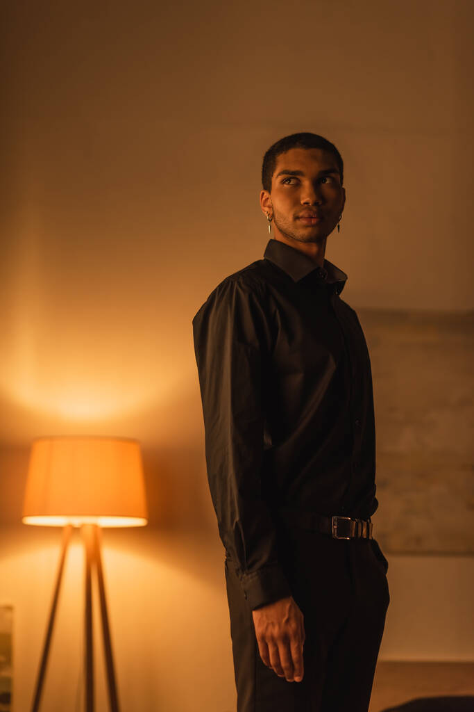 young african american man in black clothes looking away while standing in dark room with luminous lamp - Photo, image