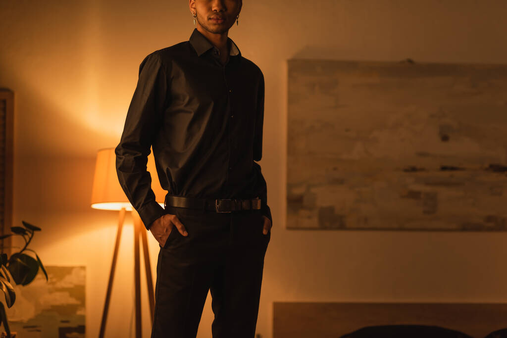 vista cortada do jovem afro-americano de pé com as mãos em bolsos de calças pretas no quarto escuro com iluminação - Foto, Imagem