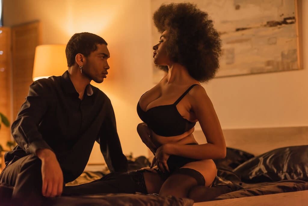 セクシーなアフリカ系アメリカ人女性で下着姿で黒シャツの若い男の近くのベッドに座って - 写真・画像