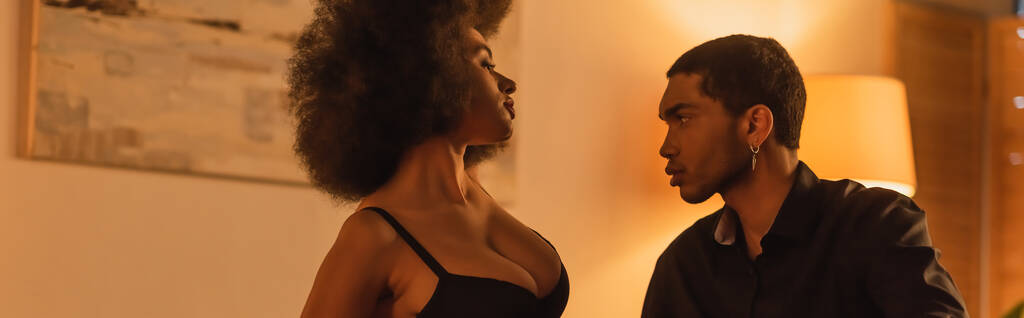 sexy donna afroamericana in reggiseno vicino giovane fidanzato in camicia nera in camera da letto a casa, banner - Foto, immagini