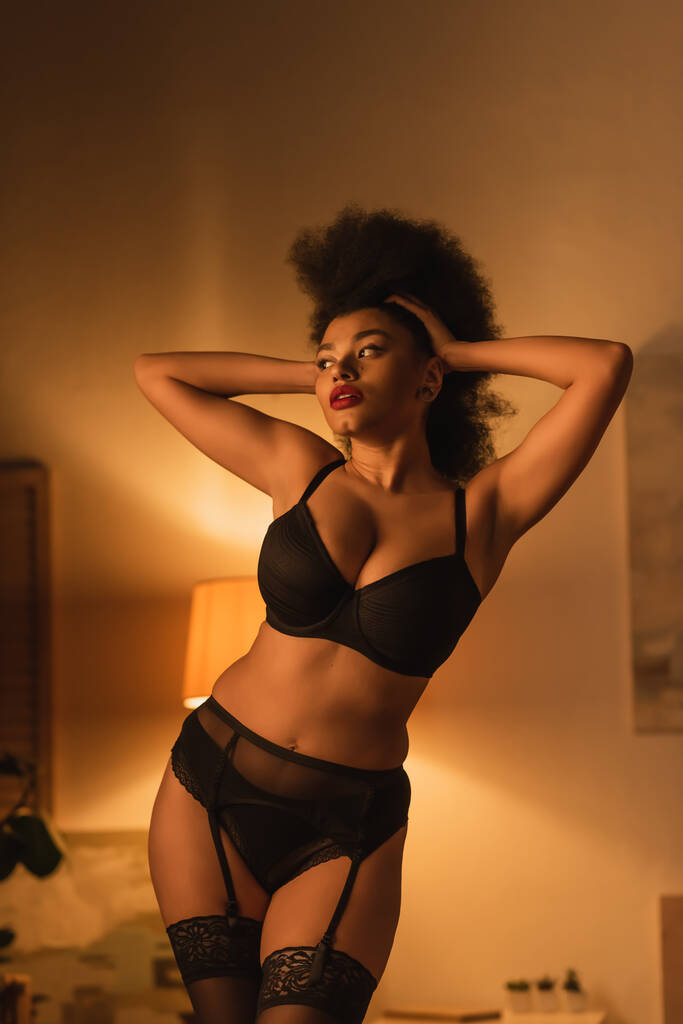 σέξι αφροαμερικανή γυναίκα με μαύρα εσώρουχα αγγίζει σγουρά μαλλιά και κοιτάζει μακριά στο σκοτεινό υπνοδωμάτιο με φωτισμό - Φωτογραφία, εικόνα