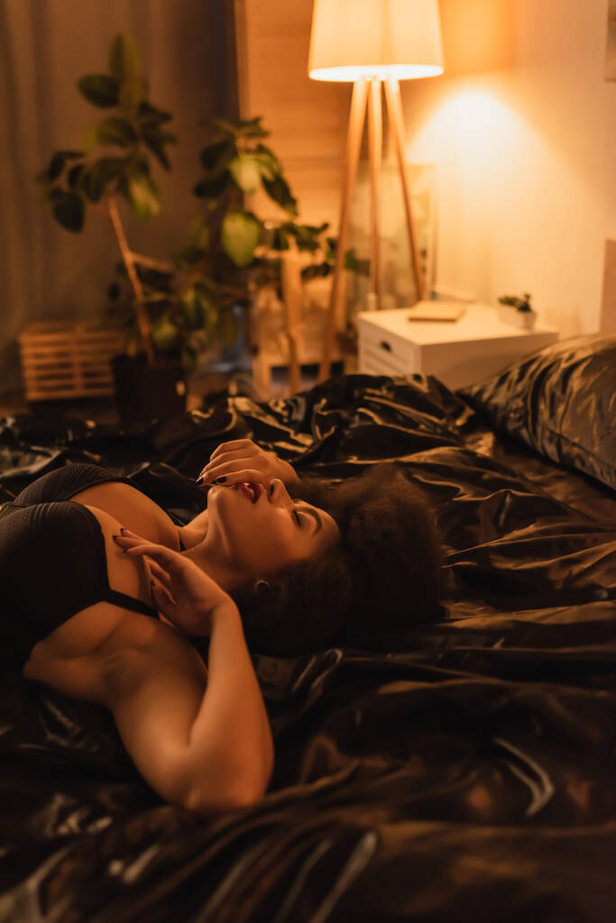 young and sexy african american woman in black bra lying on black satin bedding in dark bedroom with lighting - Φωτογραφία, εικόνα