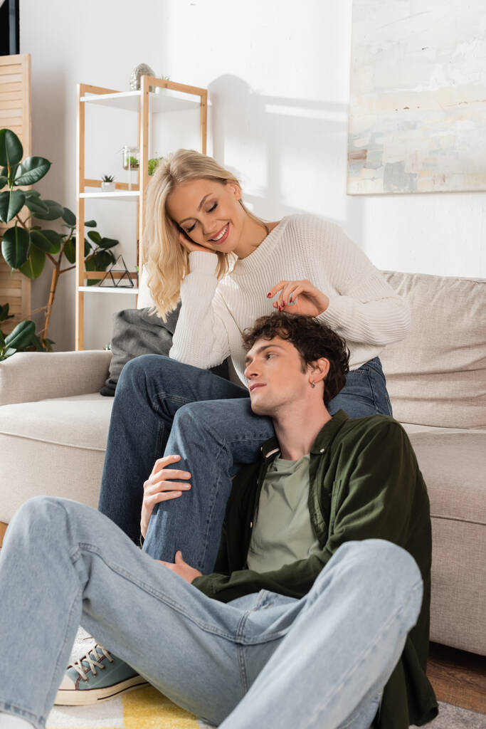 young man hugging leg of happy girlfriend in living room  - 写真・画像