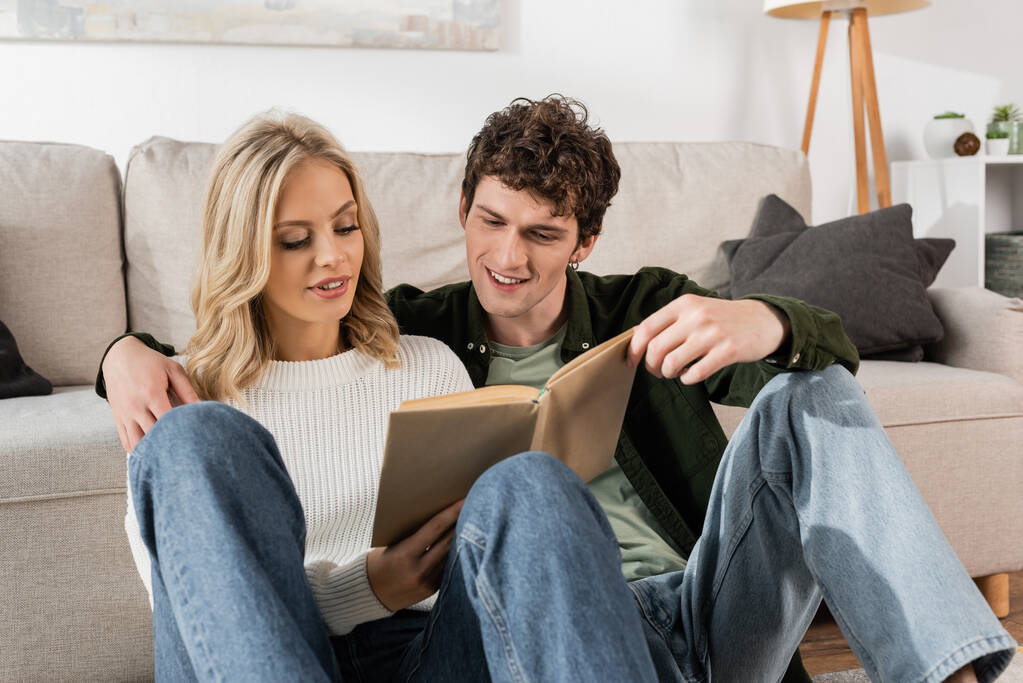 young couple smiling while reading book together in living room  - Photo, Image