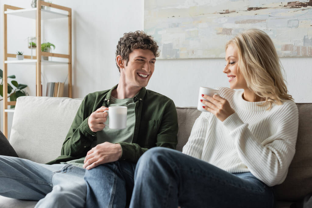 cheerful young man and blonde woman holding cups with coffee in living room  - Foto, Imagen
