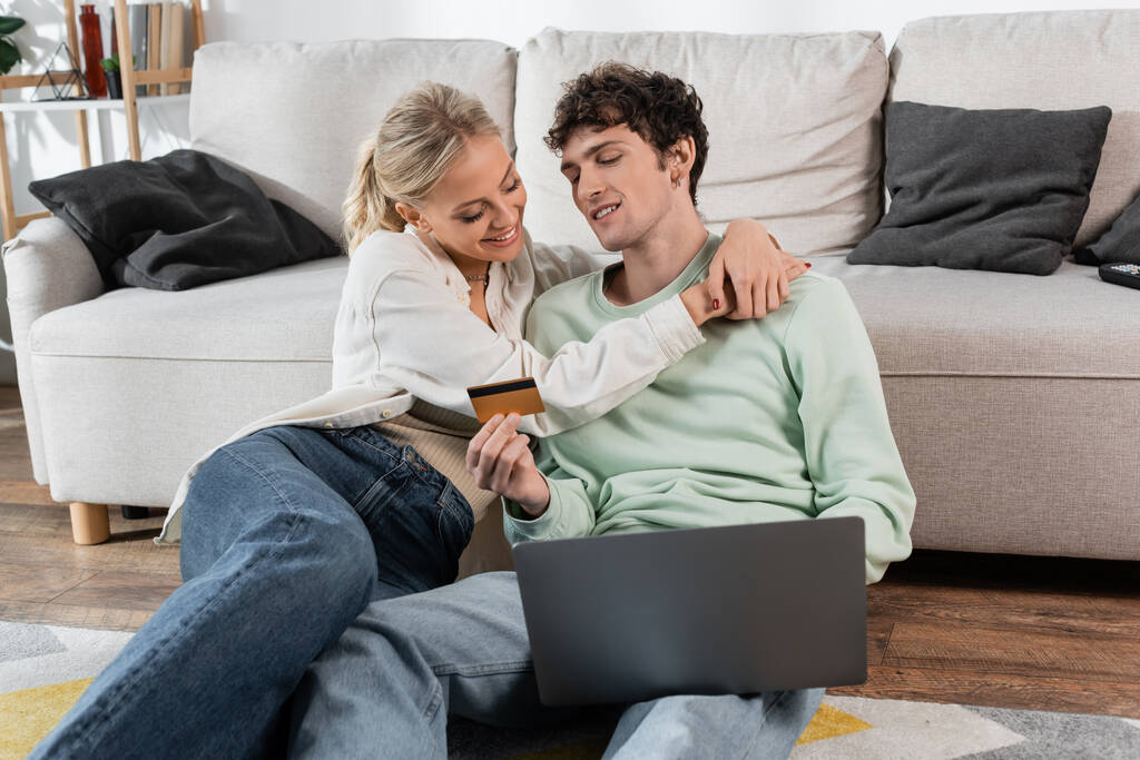 happy blonde woman hugging boyfriend holding credit card while using laptop  - Foto, afbeelding