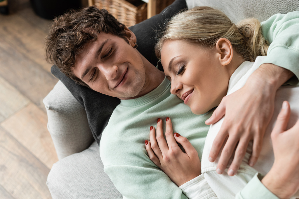 top view of happy young man embracing girlfriend while lying on couch in living room  - Zdjęcie, obraz