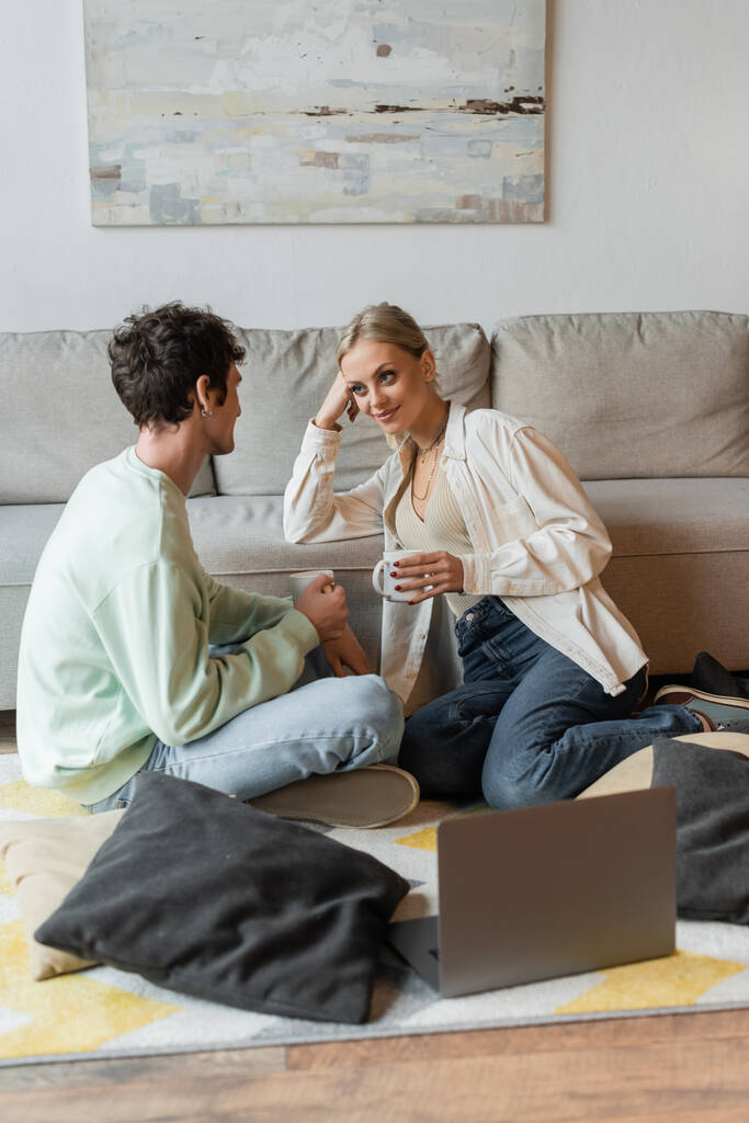cheerful young couple holding cups of coffee near pillows and laptop on carpet  - Фото, изображение