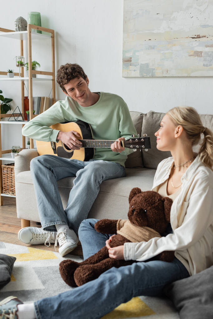 curly man sitting on couch and playing acoustic guitar near blonde woman with teddy bear  - Fotografie, Obrázek
