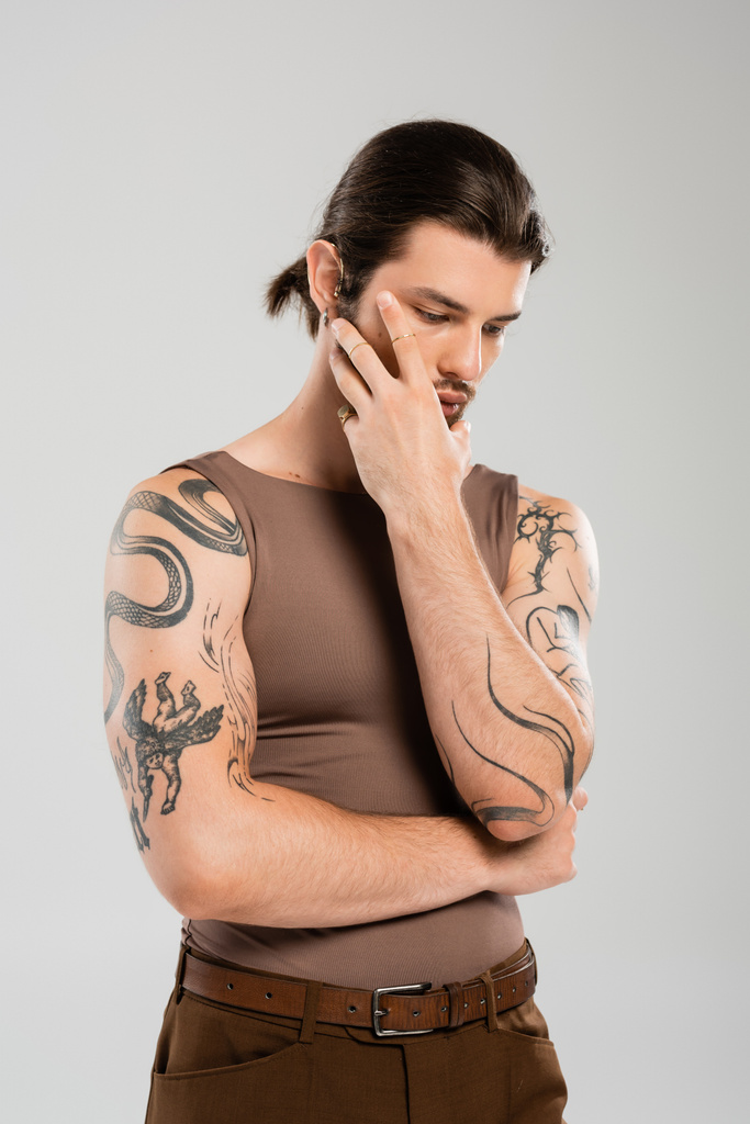 Long haired and tattooed man touching face while posing isolated on grey  - Fotó, kép