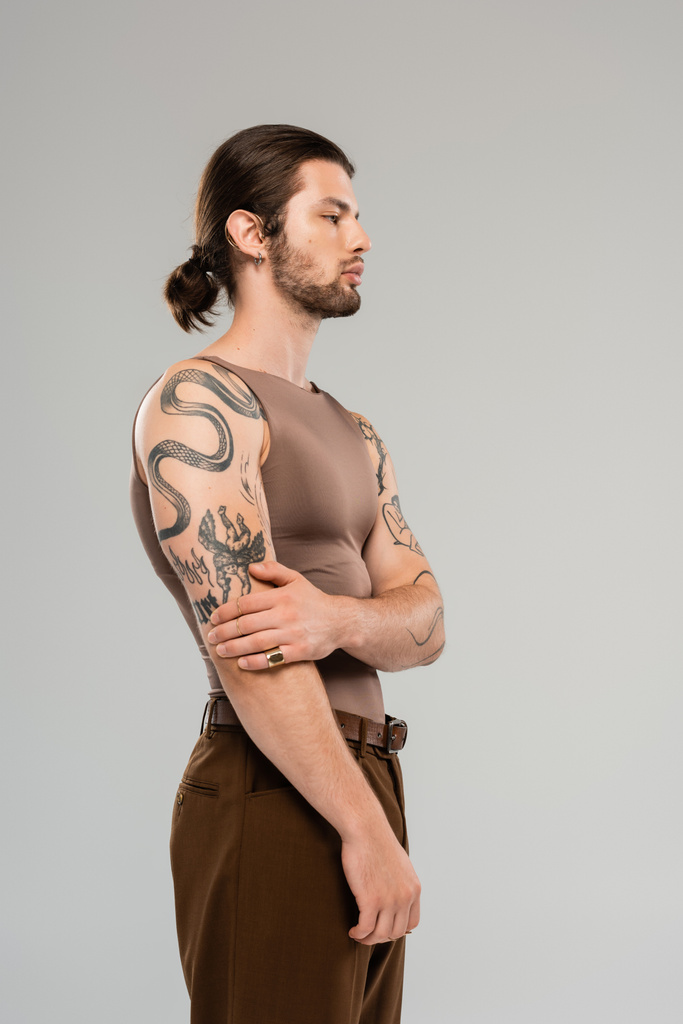 Вид збоку молодого татуйованого чоловіка в коричневому баку, що стоїть зверху ізольовано на сірому
  - Фото, зображення