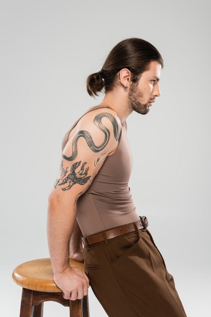 Sivunäkymä pitkäkarvainen ja tatuoitu mies poseeraa lähellä tuoli eristetty harmaa  - Valokuva, kuva