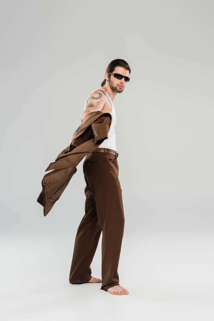 Full length of stylish tattooed man in sunglasses holding jacket on grey background - Foto, immagini