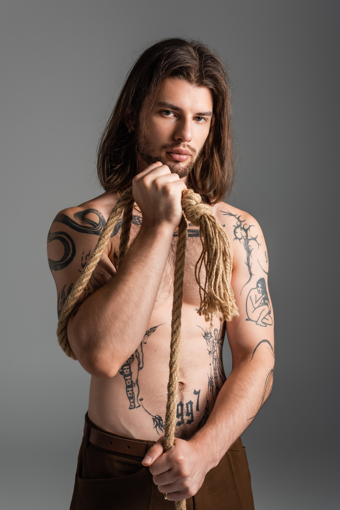 Muscular and tattooed model looking at camera while holding rope isolated on grey  - Foto, Imagem