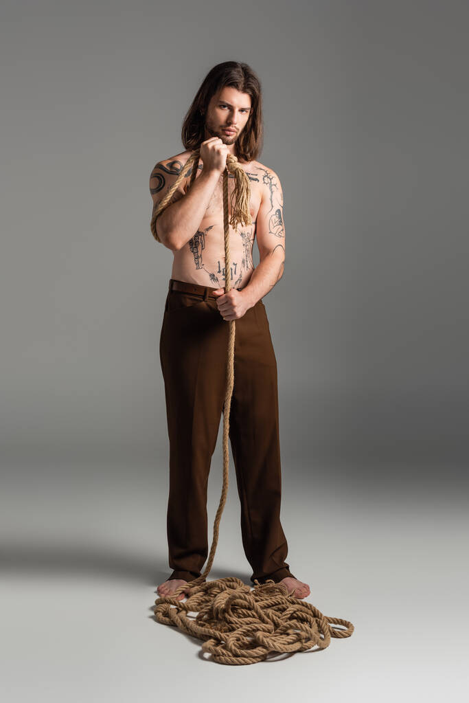 Повна довжина татуйованого чоловіка в штанях, що тримають мотузку на сірому фоні
 - Фото, зображення