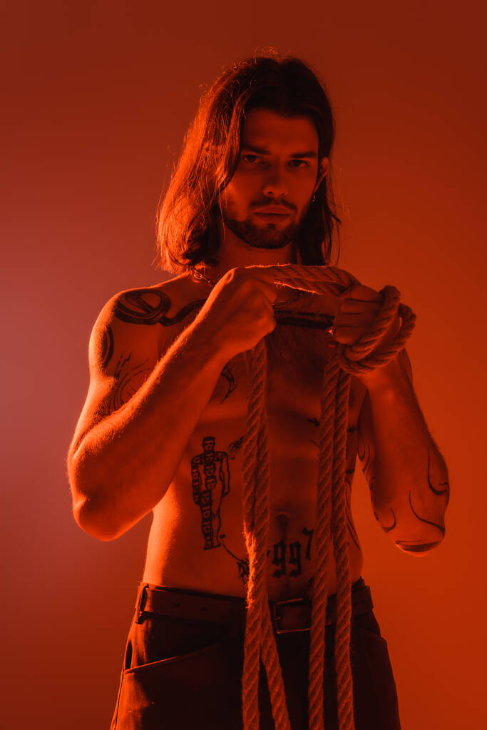 Sexy tattooed man holding rope isolated on red with light  - Foto, afbeelding