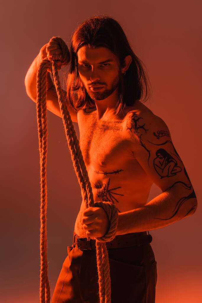 Long haired and tattooed model holding rope and looking at camera on red background  - Foto, Imagem