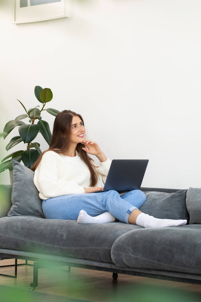 Egy vonzó barna nő laptopot használ, ül a kanapén és álmodozva mosolyog. Az online vásárlás fogalma, munka otthon. - Fotó, kép