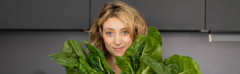 doorboorde jonge vrouw glimlachen en het houden van groene kool bladeren in de keuken, banner  - Foto, afbeelding