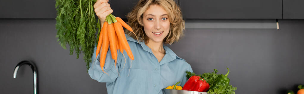 vrolijke jonge vrouw houden kom met groenten en verse wortelen in de keuken, banner  - Foto, afbeelding