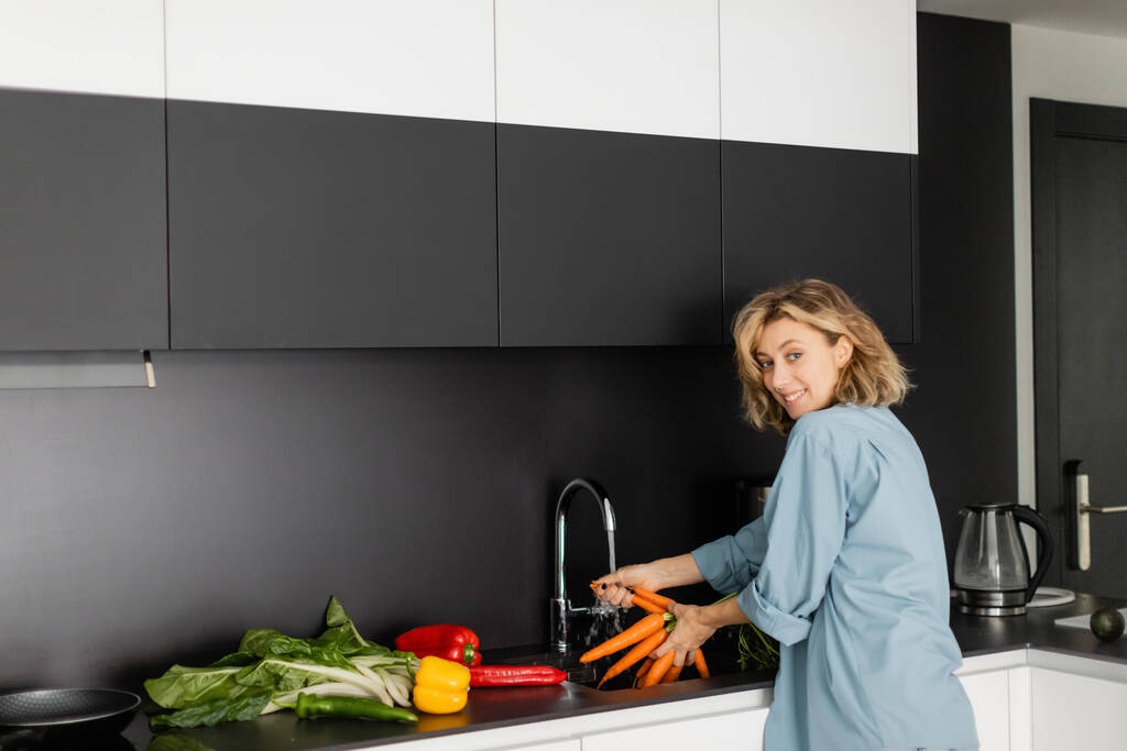 wesoła młoda kobieta myje marchewki w pobliżu świeżych warzyw w kuchni  - Zdjęcie, obraz