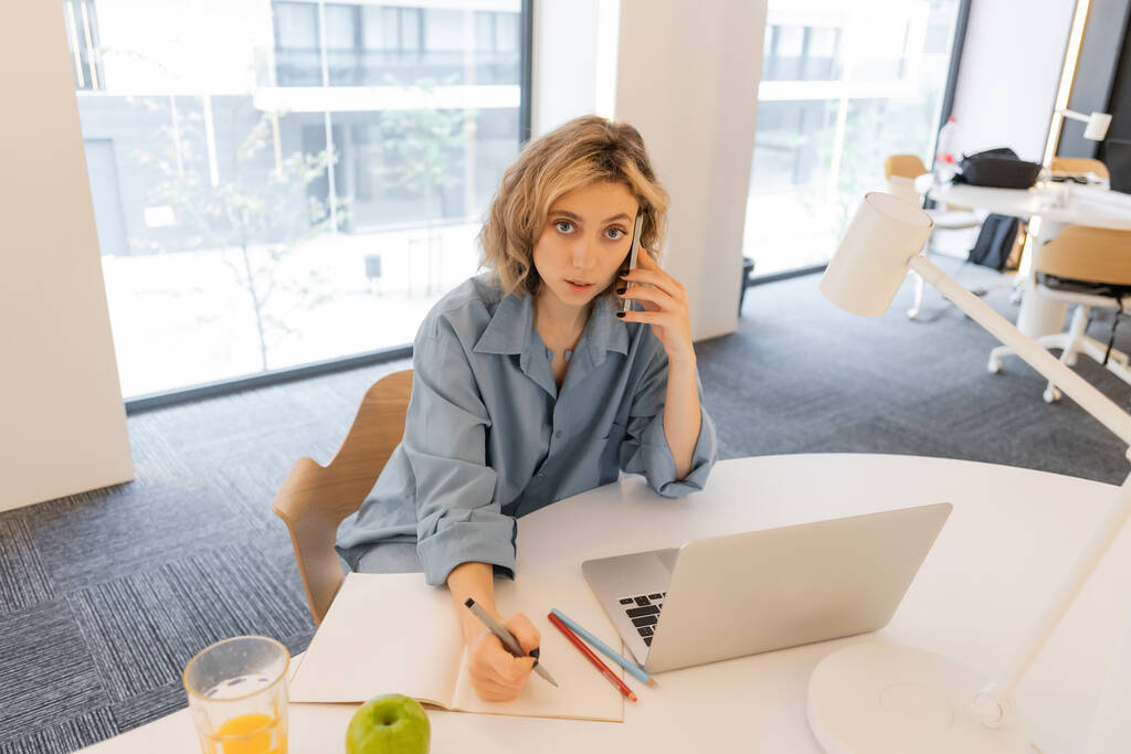 Bilgisayarın yanında akıllı telefondan konuşan genç bir kadının yüksek açılı görüntüsü.  - Fotoğraf, Görsel