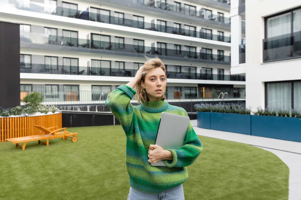 blonde woman in green sweater holding laptop and adjusting wavy hair near hotel building in Barcelona  - 写真・画像