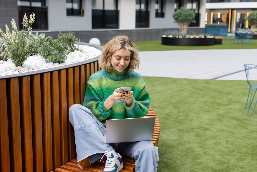 boldog fiatal nő zöld pulóverben ül fa padon laptop használata közben okostelefon  - Fotó, kép