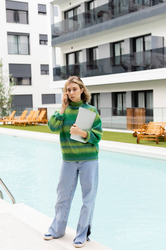 donna bionda in maglione in piedi con computer portatile mentre parla su smartphone vicino alla piscina all'aperto a Barcellona  - Foto, immagini