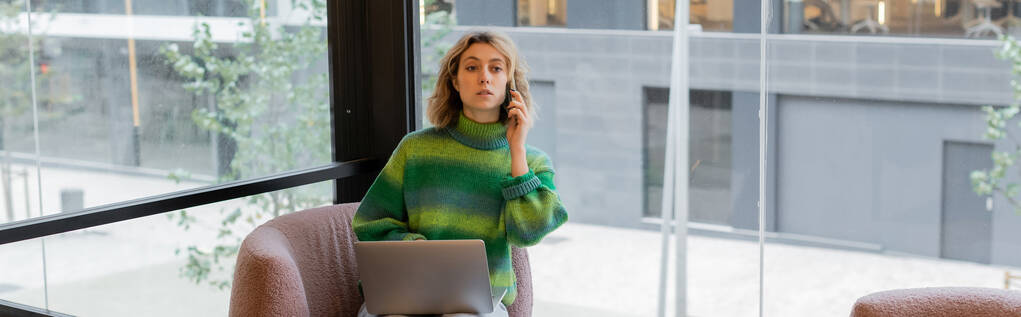 junge Frau im Pullover spricht mit Smartphone, während sie mit Laptop in der Lobby eines Hotels in Barcelona sitzt, Banner  - Foto, Bild