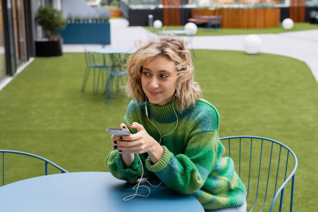 happy young woman in green sweater listening music in wired earphones and holding smartphone  - Foto, immagini