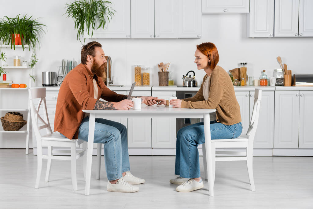 Side view of couple holding hands near laptop and cups in kitchen  - Valokuva, kuva