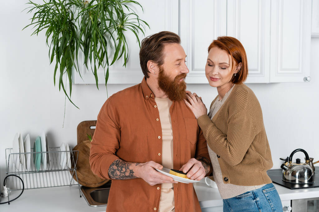 Redhead woman hugging husband washing plate in kitchen  - Fotografie, Obrázek