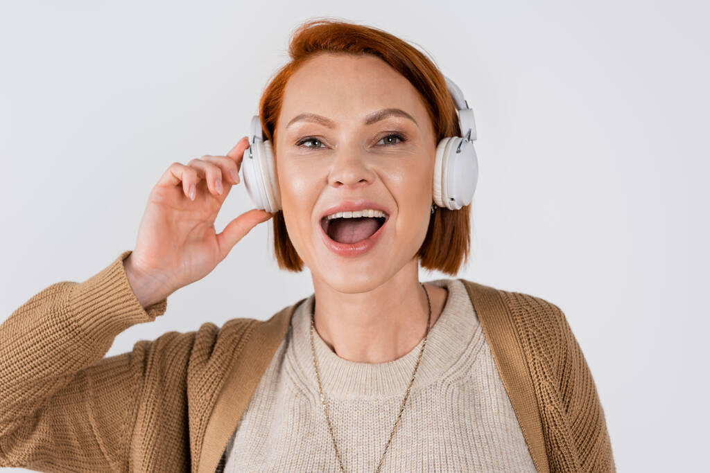 Portrait of red haired woman singing while using headphones isolated on grey  - Photo, Image