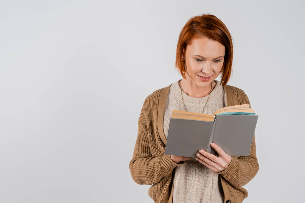 Lächelnde rothaarige Frau liest Buch isoliert auf grau  - Foto, Bild