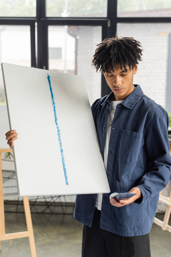 Νεαρός Αφροαμερικάνος καλλιτέχνης που κρατάει καμβά και ζωγραφίζει στο στούντιο  - Φωτογραφία, εικόνα