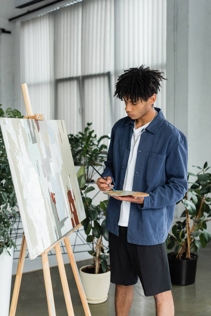 Africký americký umělec drží paletu a štětec v blízkosti kreslení na stojanu  - Fotografie, Obrázek