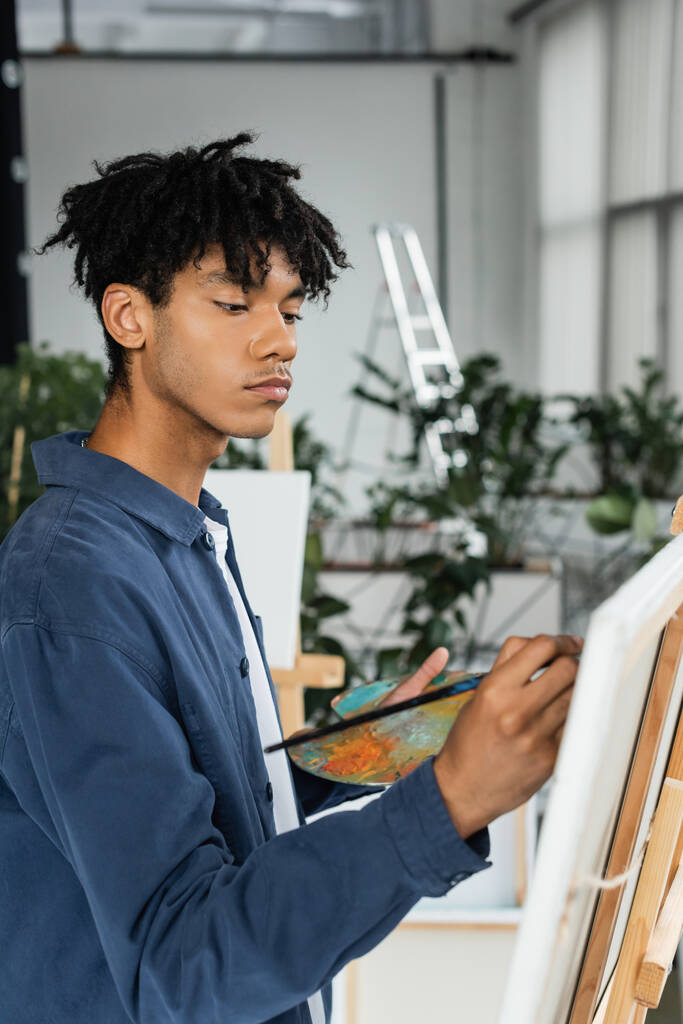 African american artist painting on blurred canvas and holding palette in studio  - Photo, Image