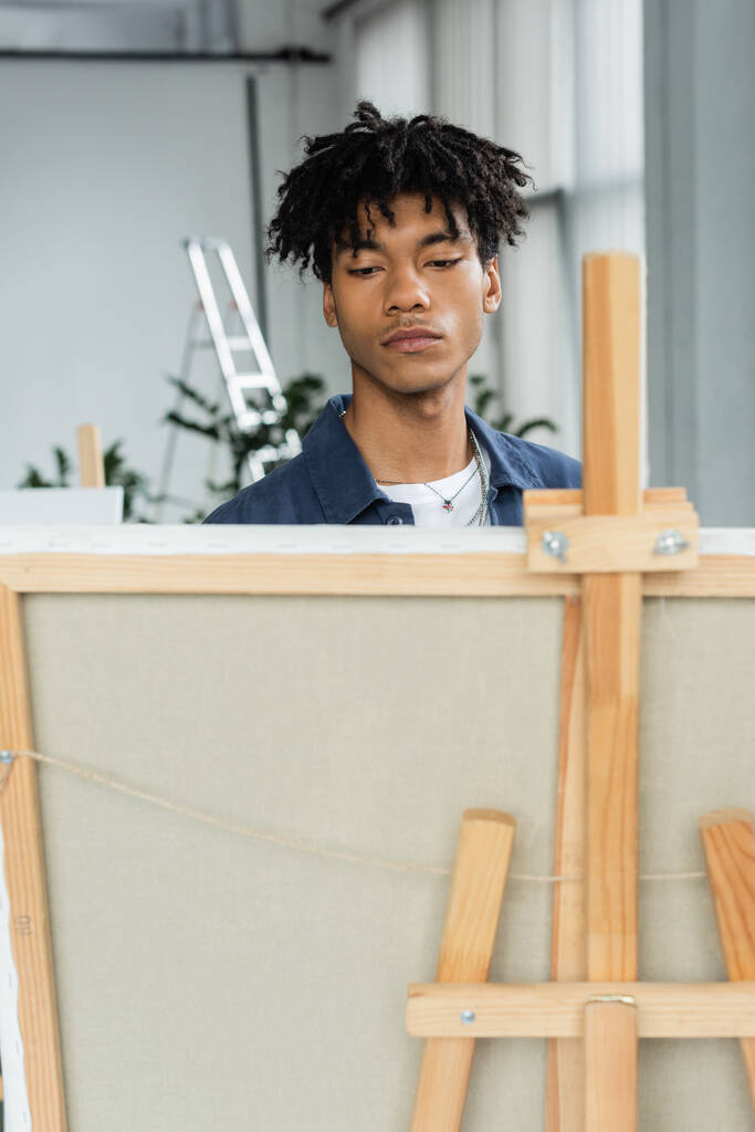 Young african american artist looking at blurred canvas on easel in workshop  - Fotografie, Obrázek