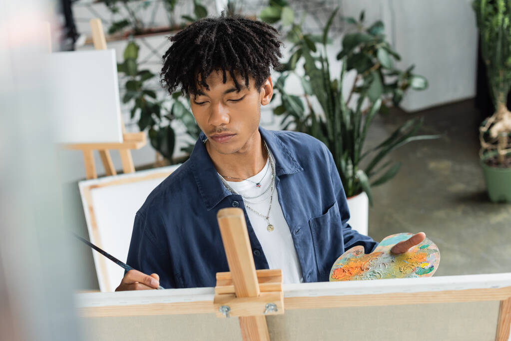 Genç Afrikalı Amerikalı ressam tuval üzerinde resim yapıyor ve paleti stüdyoda boyayla tutuyor.  - Fotoğraf, Görsel