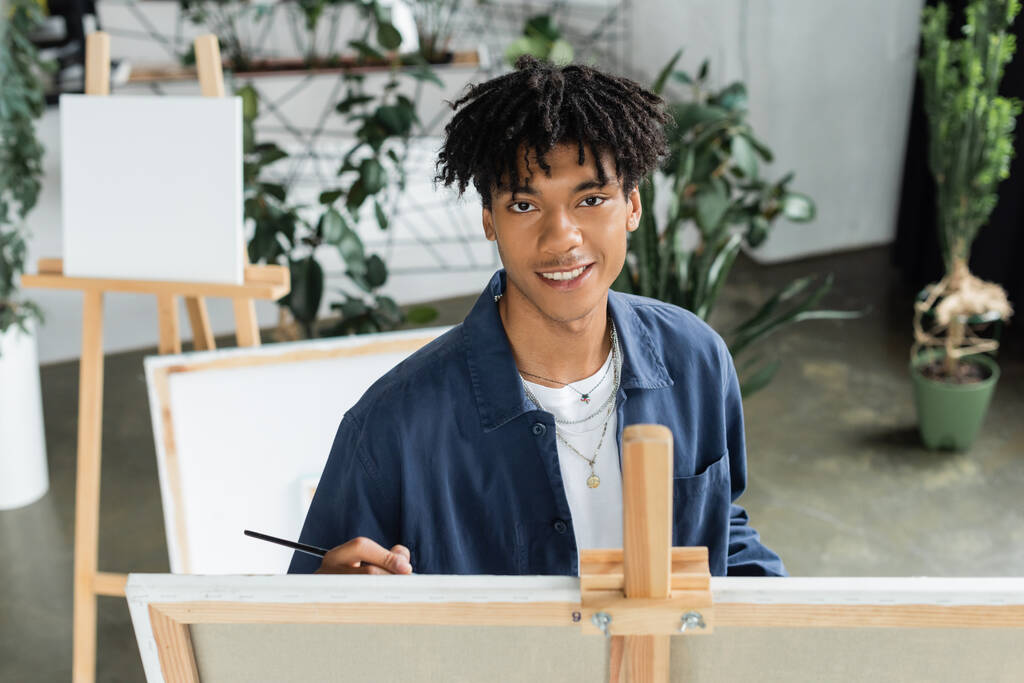 Усміхнений афроамериканський художник тримає пензлик і дивиться на камеру біля полотна в студії
  - Фото, зображення