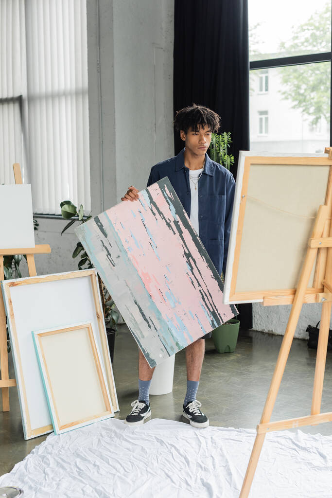 Mladý africký americký umělec drží obraz v blízkosti plátna na stojanu ve studiu  - Fotografie, Obrázek