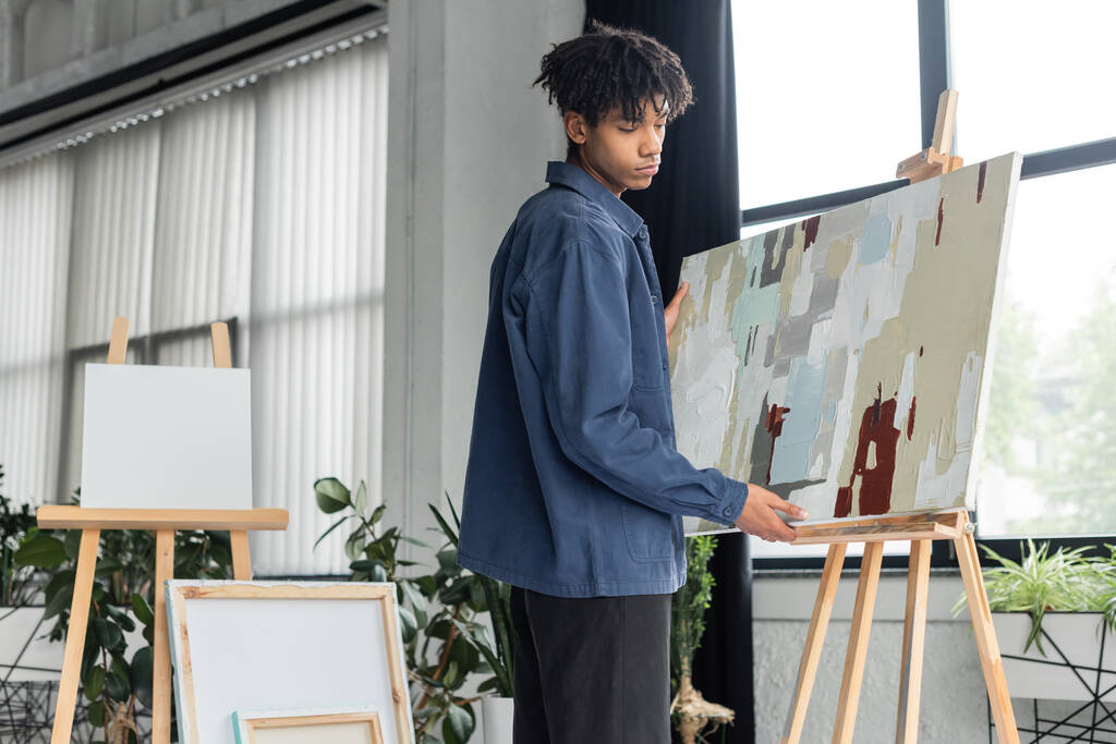 Νεαρός Αφροαμερικάνος καλλιτέχνης που παίρνει ζωγραφική από το καβαλέτο στο εργαστήριο  - Φωτογραφία, εικόνα