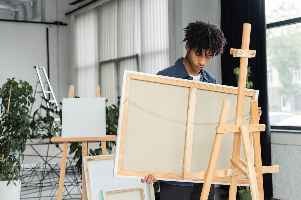 Young african american artist holding canvas near wooden easel in studio  - Foto, imagen
