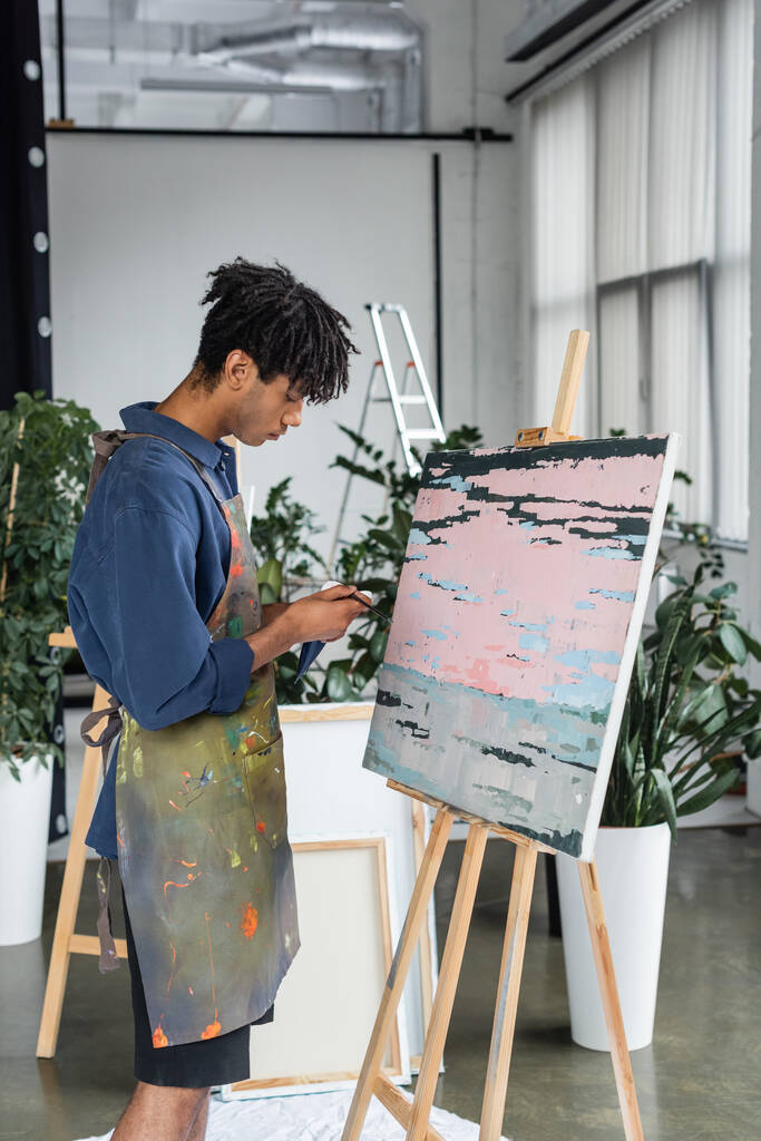 Vista lateral del artista afroamericano en delantal sosteniendo pincel y pintura cerca del dibujo en el estudio  - Foto, Imagen