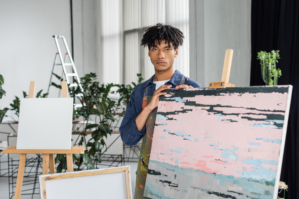 Jovem artista afro-americano em avental olhando para câmera perto de desenho em oficina  - Foto, Imagem