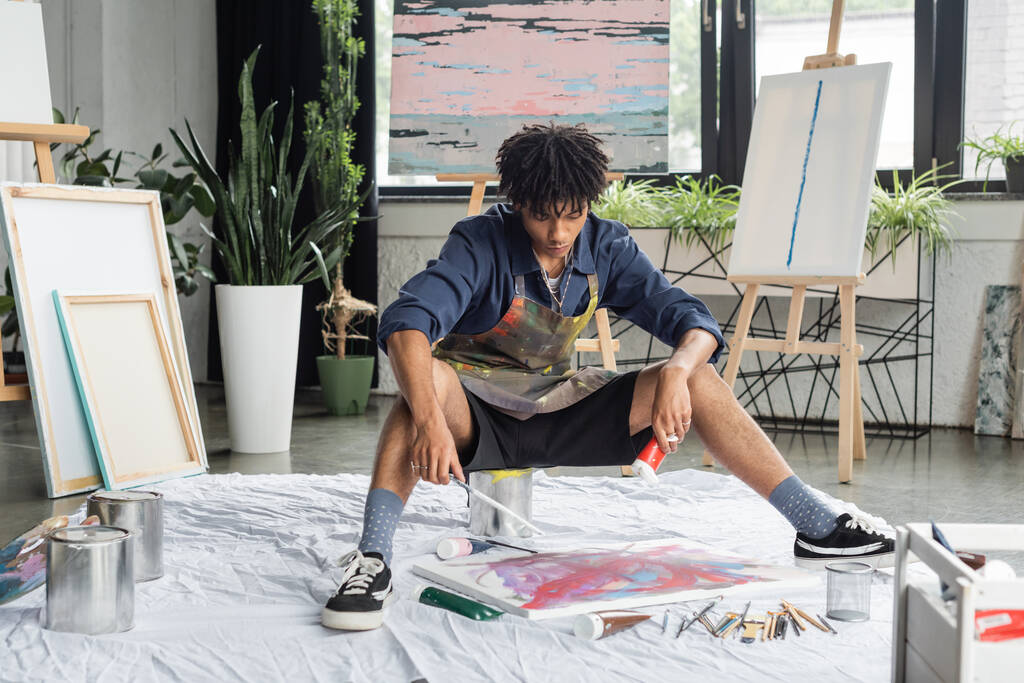 Młody afrykański artysta trzymający farbę i pędzel w pobliżu płótna na podłodze w warsztacie  - Zdjęcie, obraz