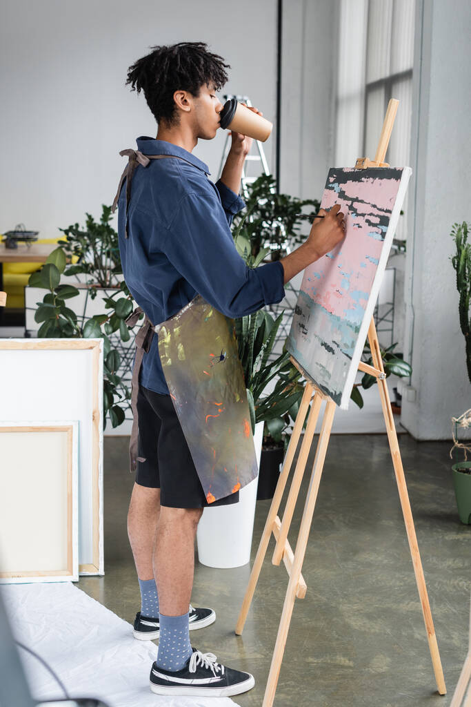 Vista lateral do jovem artista afro-americano bebendo café e pintura em tela no estúdio  - Foto, Imagem